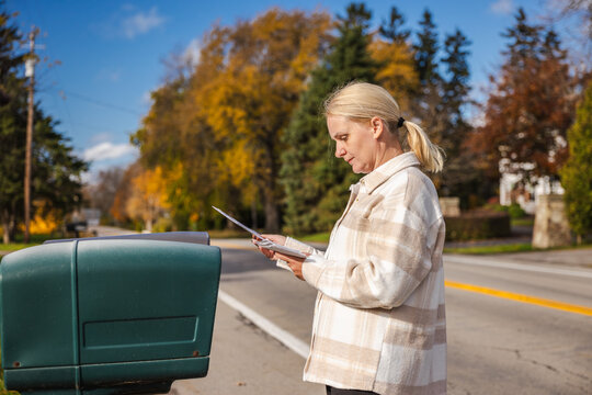 Woman standing beside a roadside mailbox on a sunny autumn day, holding an envelope and checking her mail while colorful trees line the quiet suburban street.