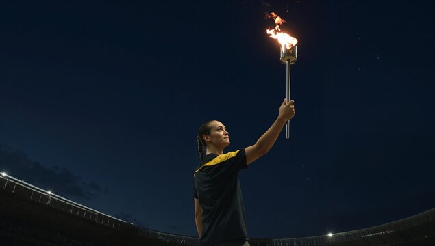 Raising athlete holding lit torch in floodlit stadium rail at night, wearing dark top yellow stripe