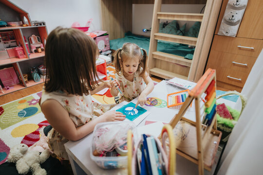 Two young girls sit together exploring arts and crafts in a cozy, colorful bedroom setting. The room is filled with toys, books, and desks fostering a joyful learning environment. - Powered by Adobe