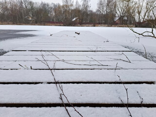 A serene winter landscape features a wooden pier, covered in fresh snow, extending over a partially frozen lake. Bare branches lie on the snow-covered planks,