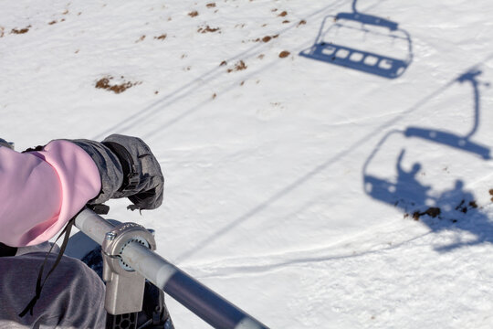 A person rides a ski lift above a snowy slope, wearing winter gloves and a pink jacket. Shadows of the lift and riders stretch across the snow, capturing a calm moment on the mountain.