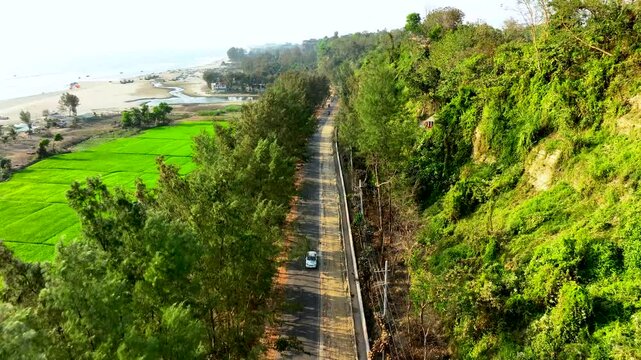 Aerial view of a coastal Merin Drive road flanked by green trees, with a beach, ocean, and a lush hillside at Cox's Bazar Bangladesh