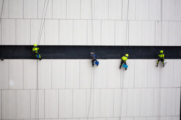 Construction crew of climbers performs dangerous task. Men in protective suits sit on seat suspended by ropes at great height. Industrial climbers work on the wall of building