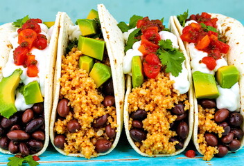 Four tacos filled with beans, seasoned meat, avocado, and salsa sit on a turquoise surface, ready to eat.