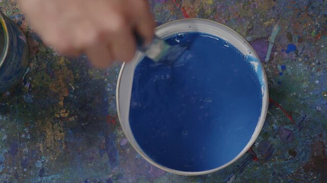 Hand r&uuml;hrt blaue Fl&uuml;ssigfarbe im Eimer um &ndash; k&uuml;nstlerischer Mischprozess mit Pigmenten, kreativer Studioumgebung und dynamischer Farbbewegung