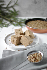 Blocks of sesame halva served on a white plate with scattered seeds and a skillet of hulled seeds in the soft, rustic kitchen scene.