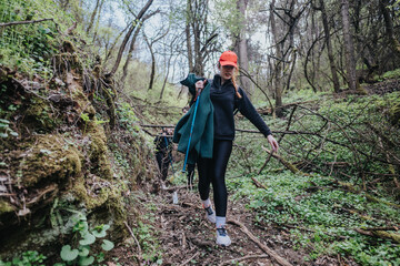 Obraz premium A woman hiker navigates a mossy forest trail, carrying a jacket and trekking poles, dressed in dark active wear and an orange cap, exploring nature and outdoor adventure.