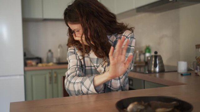 Pregnant woman in her first trimester experiencing nausea and toxicosis from the smell of cooked fish, covering her mouth and nose with her hands in the kitchen to avoid vomiting