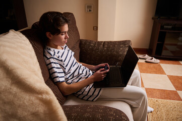 Teenage Boy Gaming At Home On Laptop In Cozy Living Room With Controller