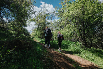 Obraz premium Two hikers trek through a lush forest on a dirt trail, carrying backpacks. Bright green trees and sunlight create a sense of outdoor adventure, fitness, and nature escape.