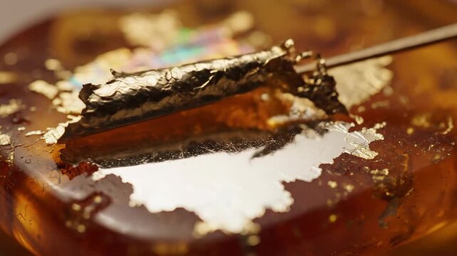 Close-up view of gold leaf being applied to a textured amber surface with iridescent reflections