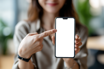 A close-up view of a woman holding a smartphone and pointing at its blank black screen in a bright, blurred office environment. The scene is ideal for showcasing app designs, user interfaces, digital 