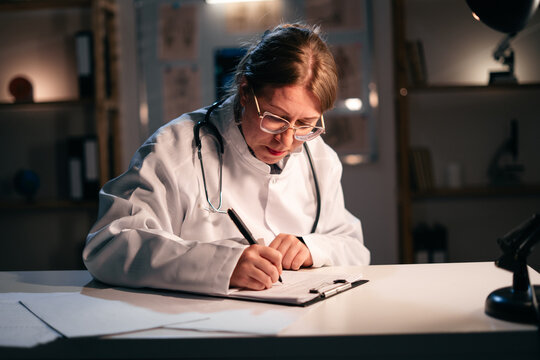 Doctor woman in white coat writing notes in documents sitting at table in clinic during night shift