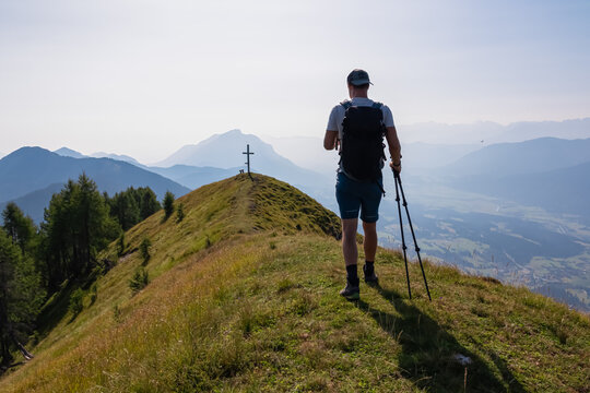 A solo male hiker with trekking poles walks along a grassy alpine ridge on Graslitzen mountain, heading towards a summit cross with a vast, hazy panoramic view of the Karawanks in Carinthia, Austria.