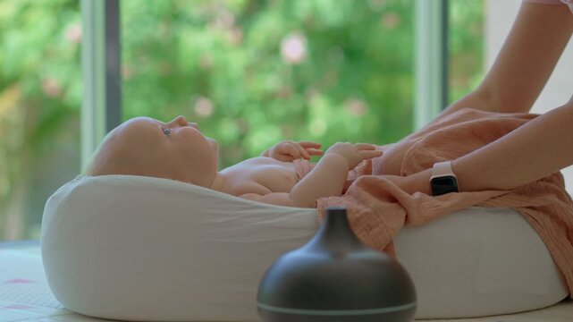 A young mother and her baby relax in a baby lounger by a large window with lush greenery outside. A working aroma diffuser humidifier adds to the calming home environment. Slowmotion video.