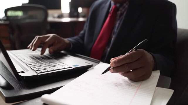 Experienced professional in a business suit multitasking, typing on laptop and writing notes in a notebook, focused on work tasks.