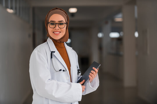 Portrait of happy Muslim female doctor or nurse wearing white coat with stethoscope in hospital corridor holding digital tablet looking at camera and smiling. Professional medical staff headshot.
