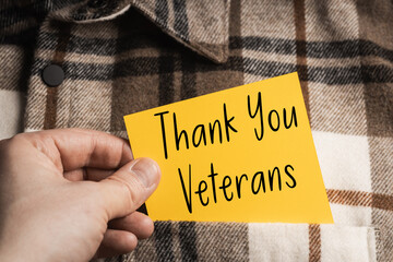 Thank You Veterans - a yellow note held in the hand against the background of a brown and beige plaid shirt, symbolizing gratitude and support for military service.