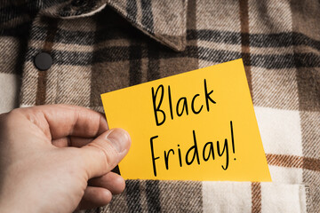 Saving on Occasions: Hand holding a bright yellow note with the inscription "Black Friday!" Against the background of a fashionable autumn shirt with a brown and beige check.
