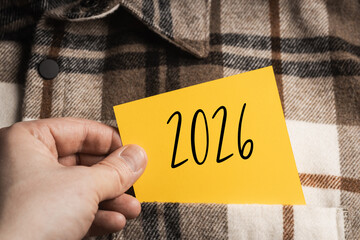 Plans for the Future: A hand holding an orange and yellow note with the date "2026" against the background of a casual shirt with a brown and beige check, symbolizing goals for the new year.