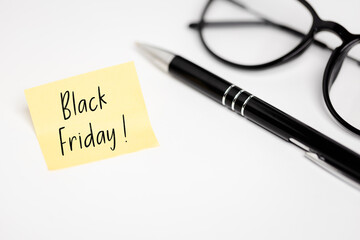 Office Shopping Planning: Yellow sticky note with the inscription "Black Friday!" Lying on a white desk next to an elegant pen and reading glasses.