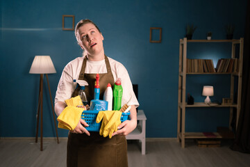 Portrait of woman in apron holding bucket with cleaning supplies making omg facial expression with...