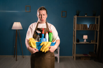 Portrait of unhappy dissatisfied woman in apron holding bucket with cleaning supplies folded look...
