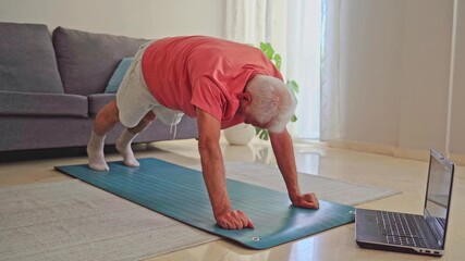 Senior man doing plank exercise at home while following online training on laptop