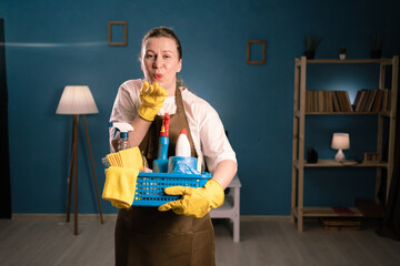 Portrait of woman in apron holding bucket with cleaning supplies sending air kiss at camera...