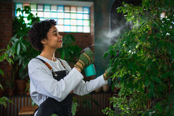 Running of own business. African woman florist with spray bottle watering fertilizing plants in botanical store, Small business owner working at flower shop smiling surrounded by plants Small business