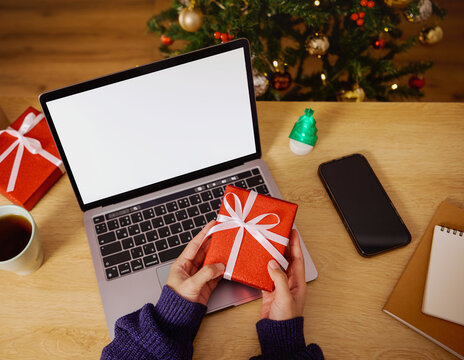 Christmas gift preparation with hands holding red present box on workspace featuring laptop and holiday decorations creating warm festive atmosphere for seasonal celebration concept
