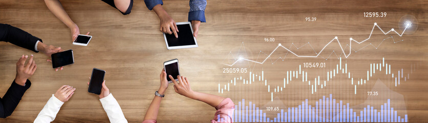 Diverse hands engaging with digital devices on a wooden surface while reviewing financial charts...
