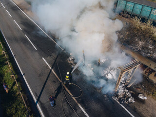 Emergency response: Firefighters in protective gear extinguish a large fire using a water hose. A high-angle shot shows the scale of the incident near transport infrastructure.