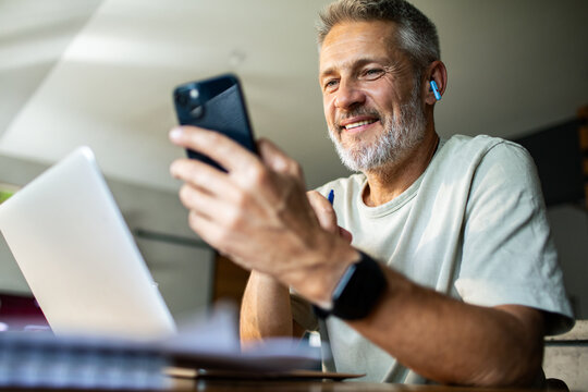 Mature man smiling using smartphone with earbuds at home office