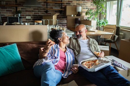 Mature married couple enjoying pizza and laughing on sofa at new home - Powered by Adobe