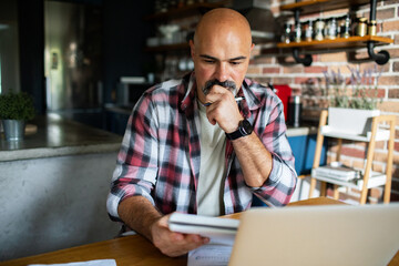 Mature man concentrating on laptop and bills in home kitchen
