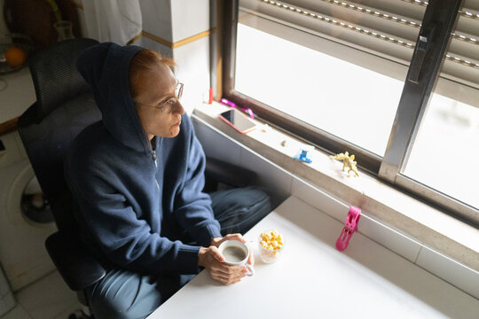 Woman drinking coffee looking out window feeling loneliness