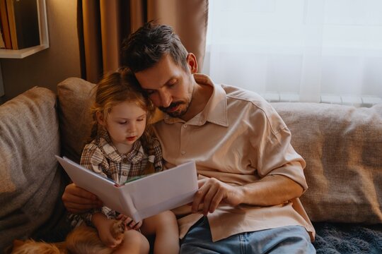 A young father comforts his 4-year-old daughter while reading her a book, sharing a tender moment of paternal love and emotional connection
