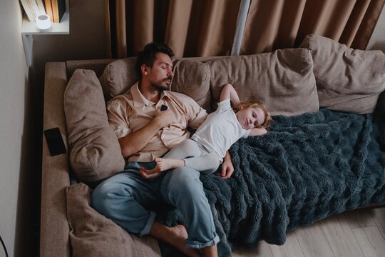 A tired young father and his 4-year-old daughter watch TV together in the evening. Authentic moment of exhausted parenting and quiet family bonding