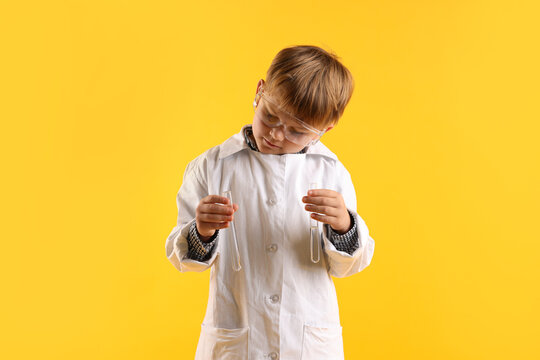 Cute little boy in laboratory coat and protective goggles with test tubes on yellow background. Child and science
