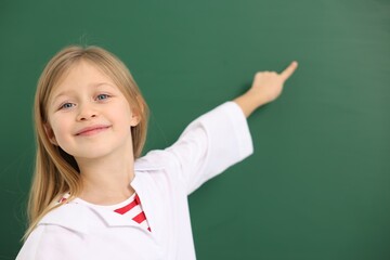 Cute little girl in laboratory coat pointing at something at green chalkboard, space for text....