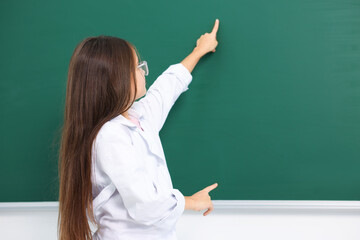 Little girl in laboratory coat and glasses pointing at something at green chalkboard indoors, space...