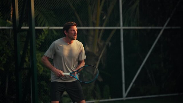 A focused adult man engages in an intense tennis match, running and hitting forehand shots on a green outdoor court. Shows dedication and athletic pursuit during evening recreation and sports.
