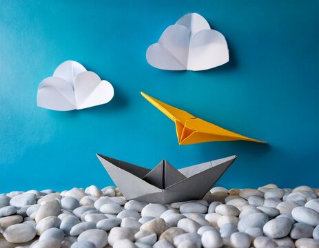 a grey origami boat rests on a surface near white pebbles a yellow paper airplane flies above it accompanied by small white origami clouds - Powered by Adobe