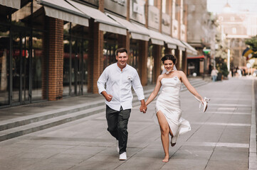 Wedding photography, portrait of happy smiling newlyweds, young groom and beautiful bride, walking, running barefoot with shoes in hands, holding hands, outdoors on the street in the city.