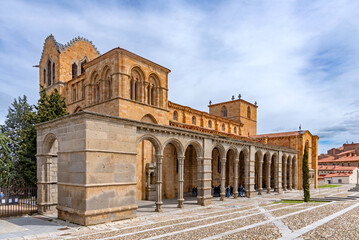 Fototapeta premium A large Romanesque church in Ávila, featuring a long arcaded portico with rounded arches and a warm golden stone exterior, standing prominently beneath a cloudy sky