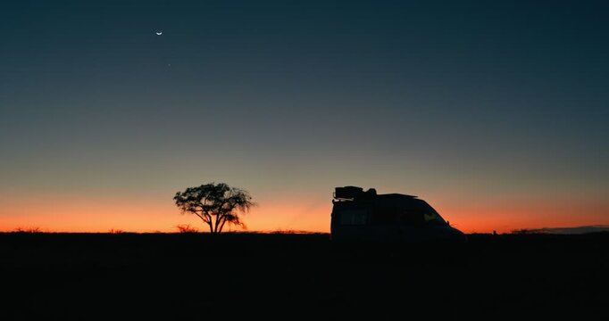Wide-angle sunrise in Namibia showing a tree and camper van silhouettes against a vivid blue-orange horizon, capturing adventure, freedom, and a cinematic desert travel atmosphere in premium quality.