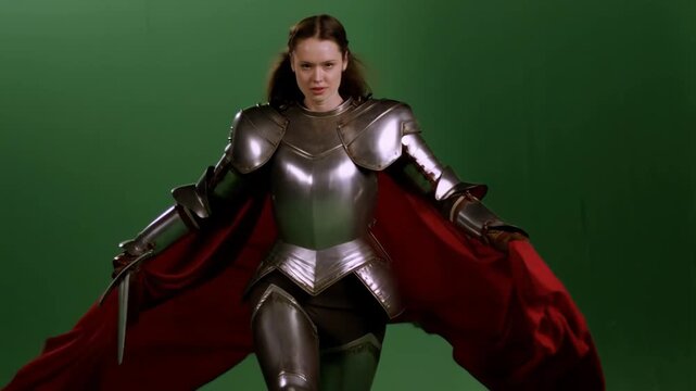 Medieval female knight in full armor with crimson cape walking against green screen, sword in hand, heroic and confident stance video footage