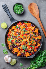 Overhead view of a fictional Southwest Sweet Potato, Black Bean and Rice Skillet on a grey slate background, with a wooden spoon, garlic, lemons and herbs.  