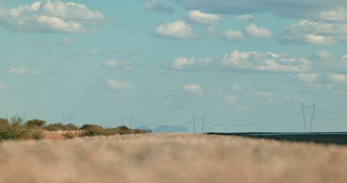 Wide shot of a desert road shimmering in the scorching heat, small clouds in a blue sky, with a distant power line and mirage-like reflection of an oncoming car on the horizon.